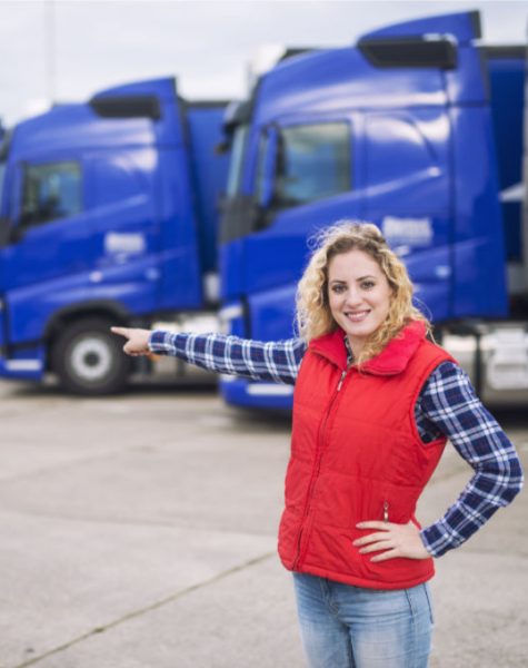 Female trucker standing in front of parked trucks and pointing her finger to the transportation vehicles. Transportation service.